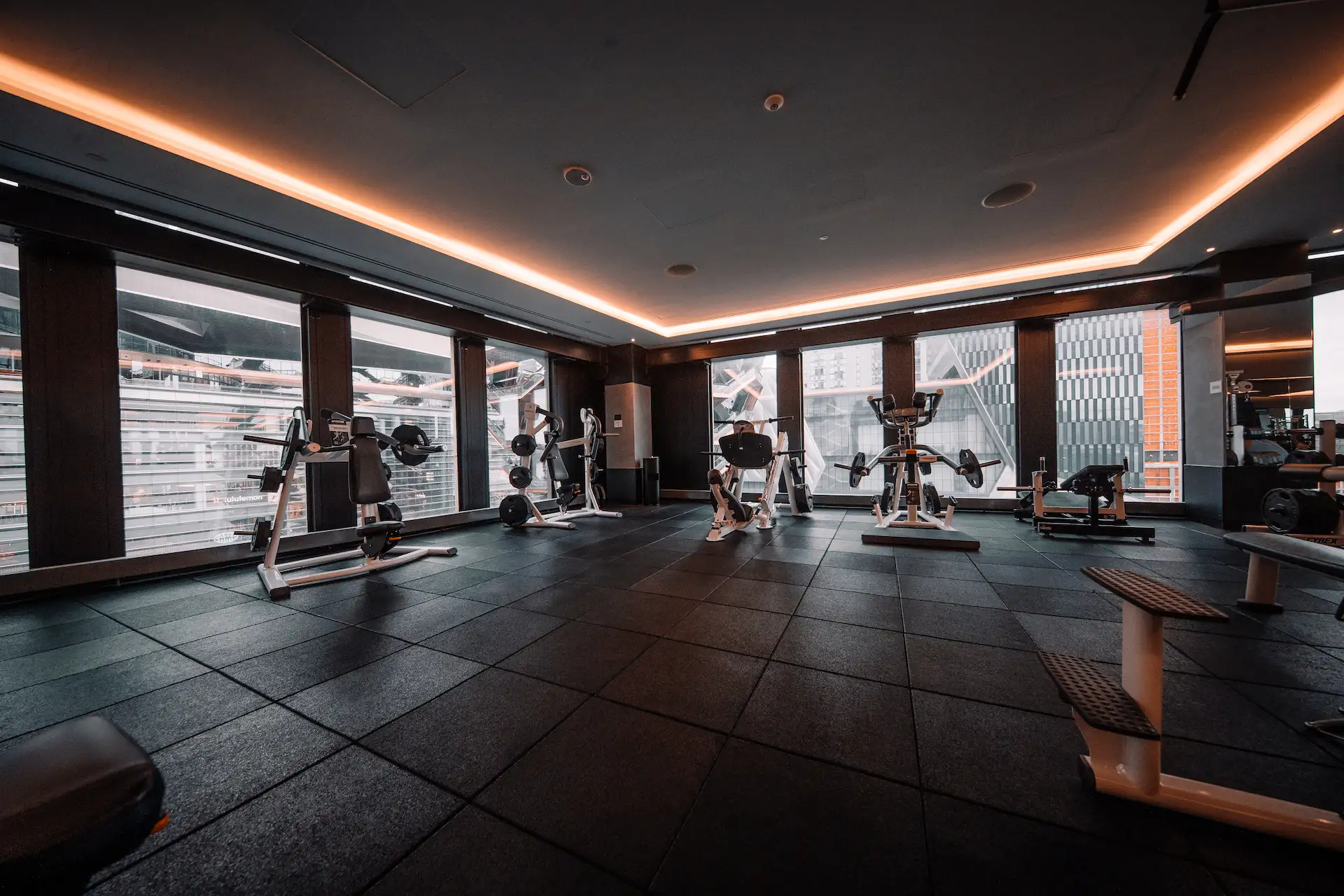 Modern indoor gym with strength training equipment, dark flooring, and floor-to-ceiling windows, evoking elevated fitness and sleek urban design