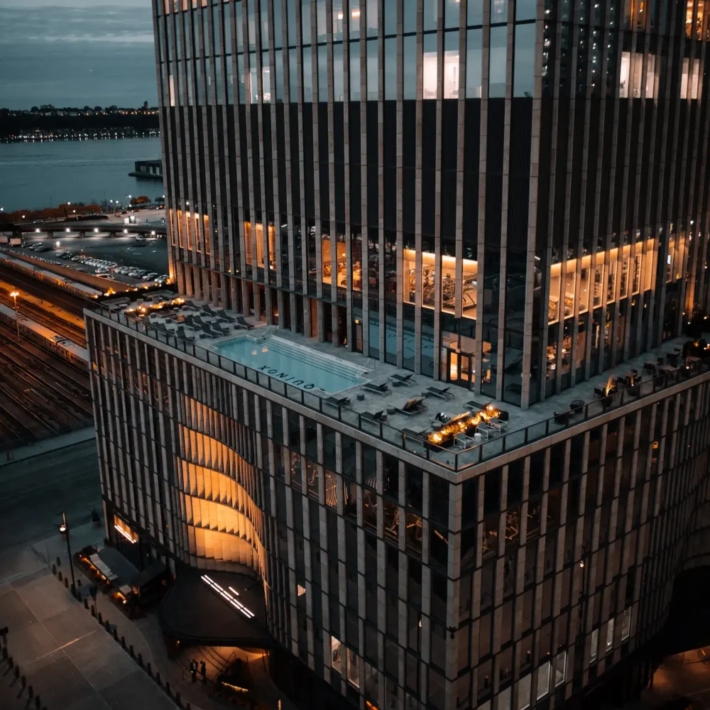 Modern high-rise building with illuminated terrace and glass facade, set against evening sky and urban waterfront, evoking upscale city living and architectural elegance
