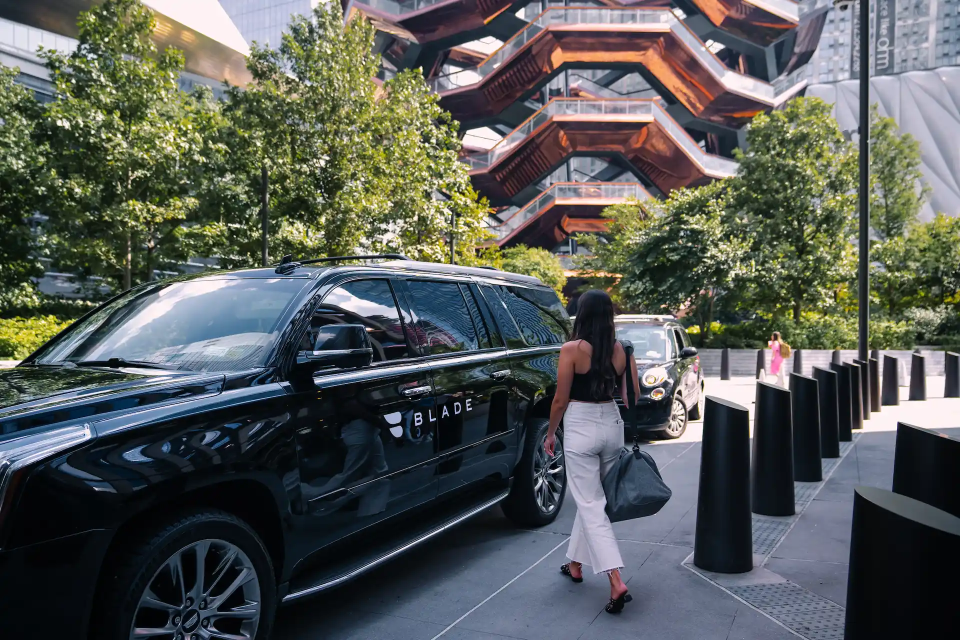 Woman walking toward black SUV branded “BLADE” near the Vessel in NYC, with modern buildings and bollards framing the upscale city scene
