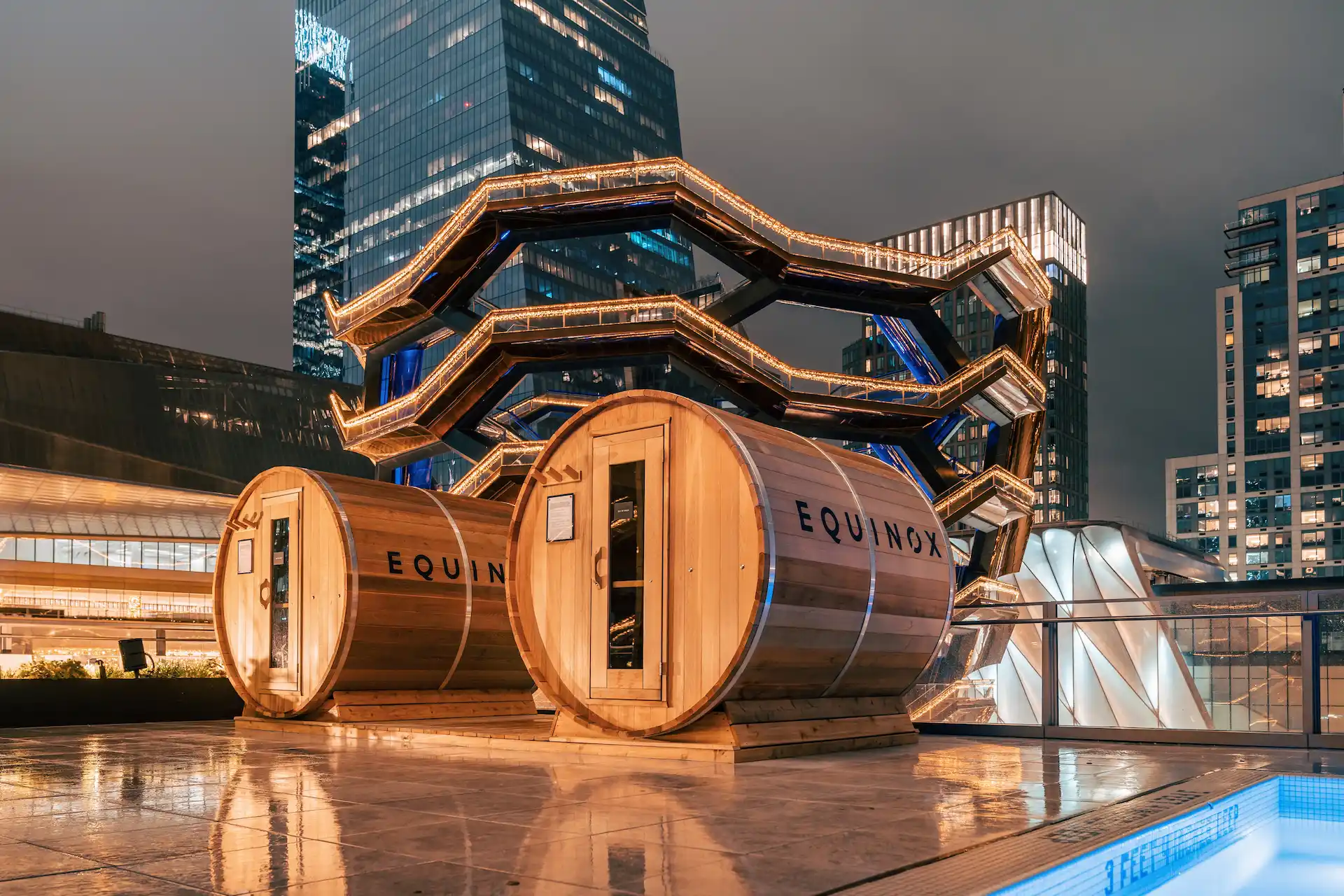 Two wooden Equinox sauna pods on rooftop at night, with reflections on wet deck and illuminated Vessel in Hudson Yards