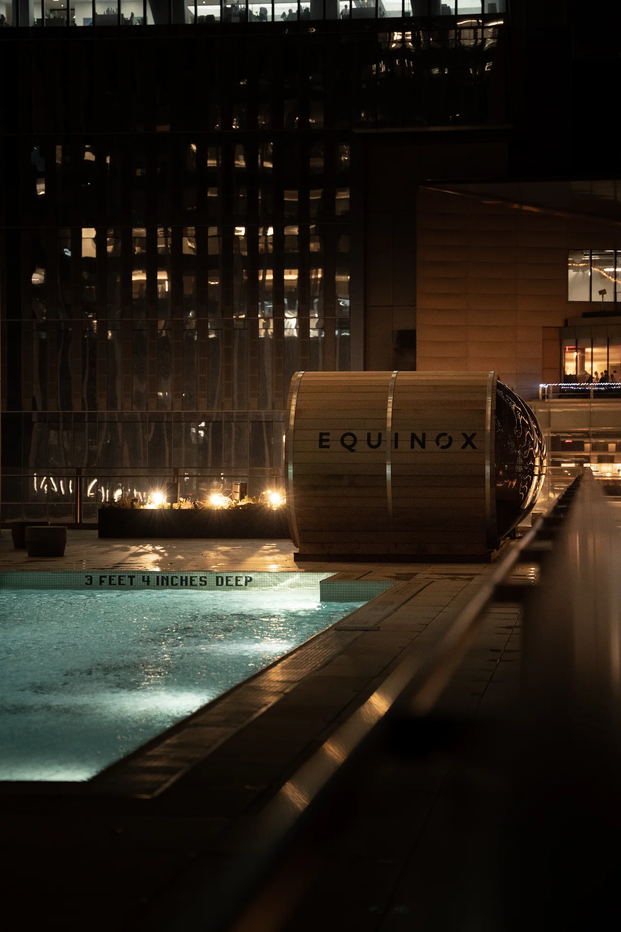 Lit rooftop pool at night beside cylindrical Equinox sauna, with glass building