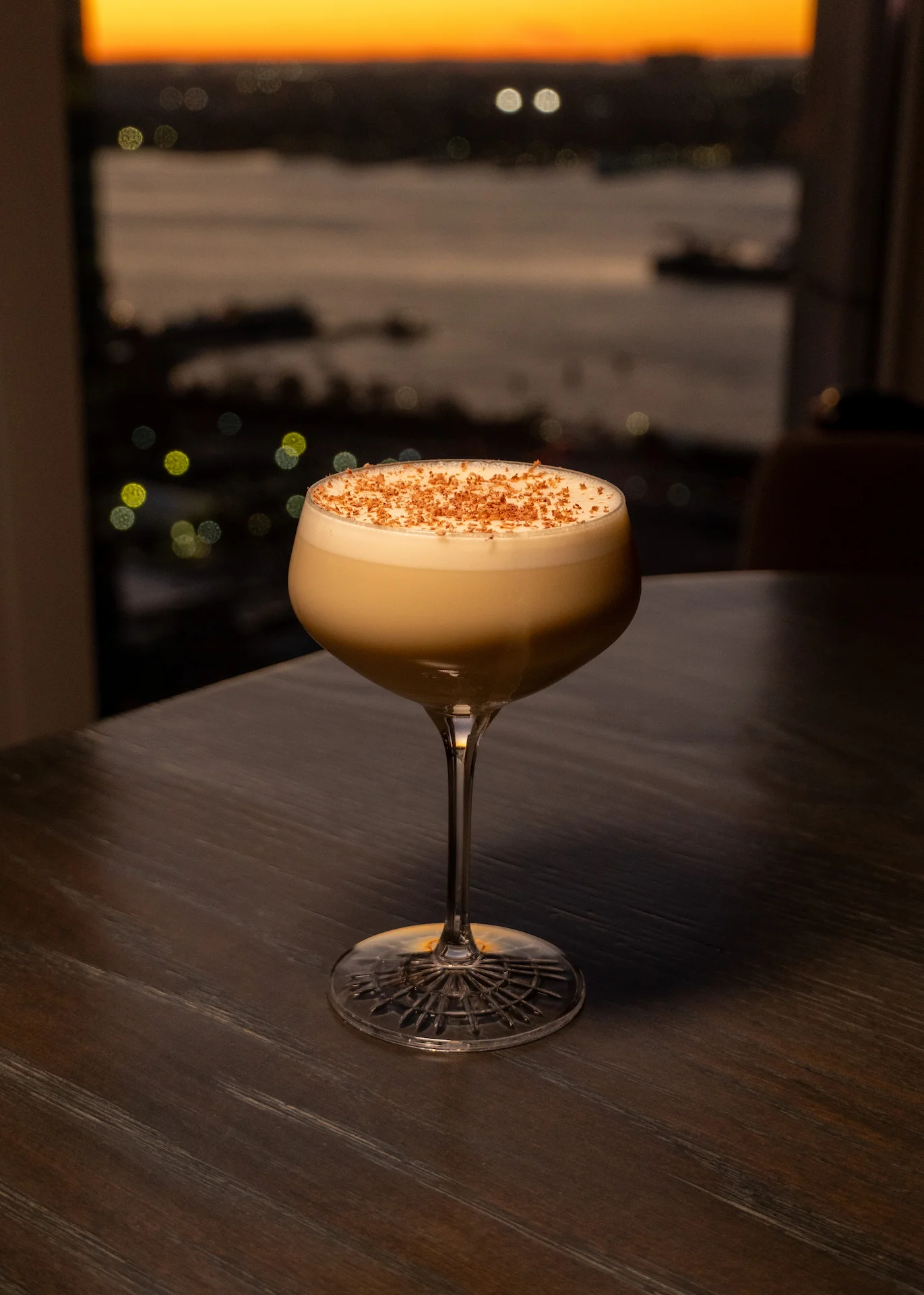 Creamy cocktail on dark table with sunset city view over water, evoking quiet indulgence