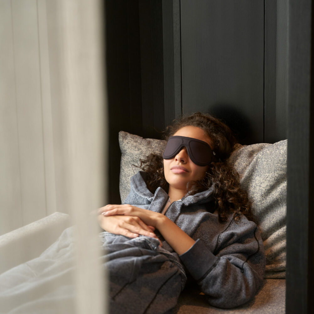Person lying down wearing sleep mask and hoodie, resting peacefully in a dim, quiet room.