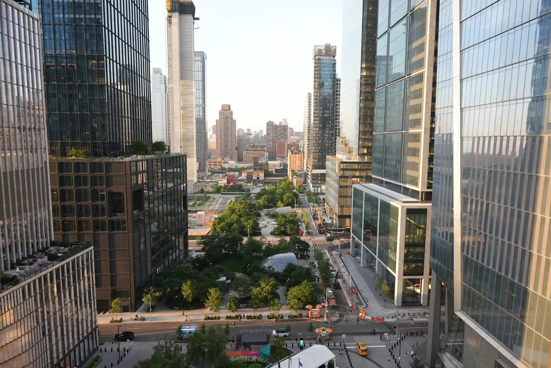 Cityscape with glass skyscrapers flanking a central green park, viewed from above with pedestrians and traffic animating the urban scene