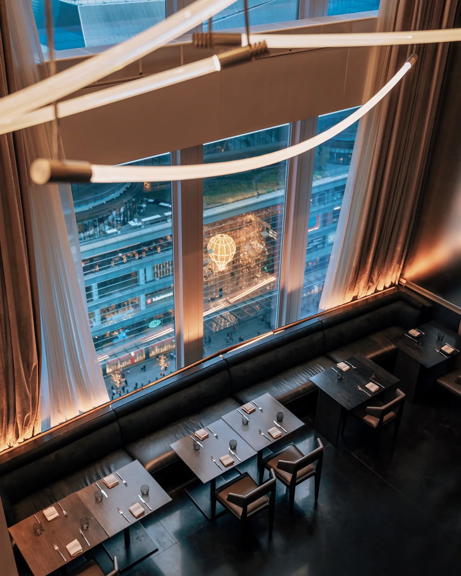 Upscale dining room with sleek dark furniture and sculptural lighting, overlooking vibrant city street through floor-to-ceiling windows