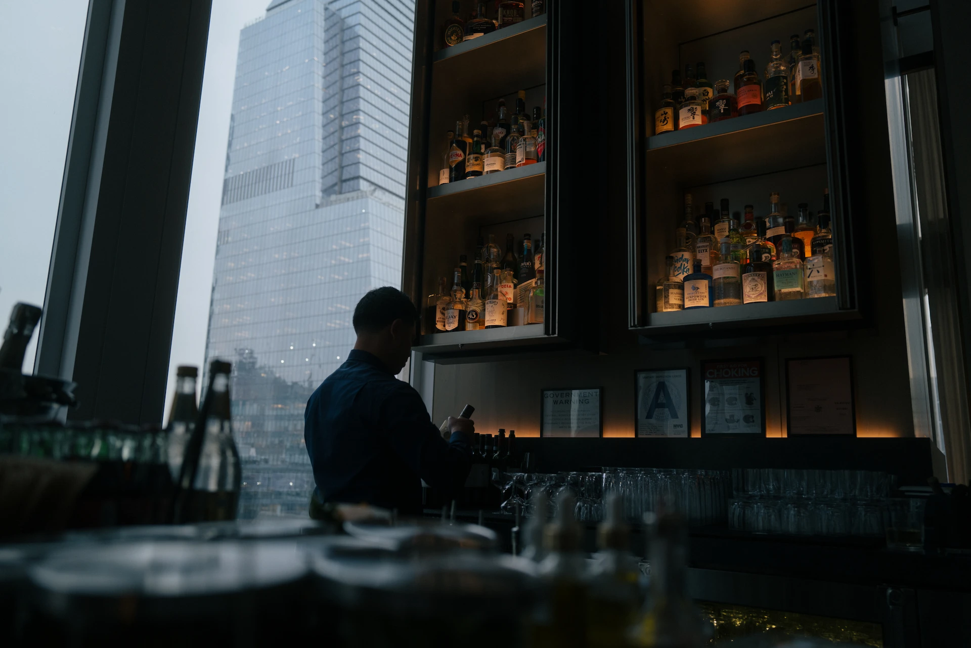 Bartender at backlit bar with liquor shelves and city view, evoking urban lounge atmosphere