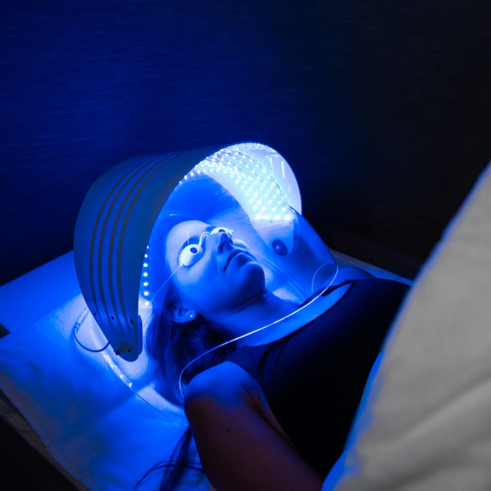 Person receiving LED light therapy, lying under curved blue light panel with protective goggles in spa-like setting.