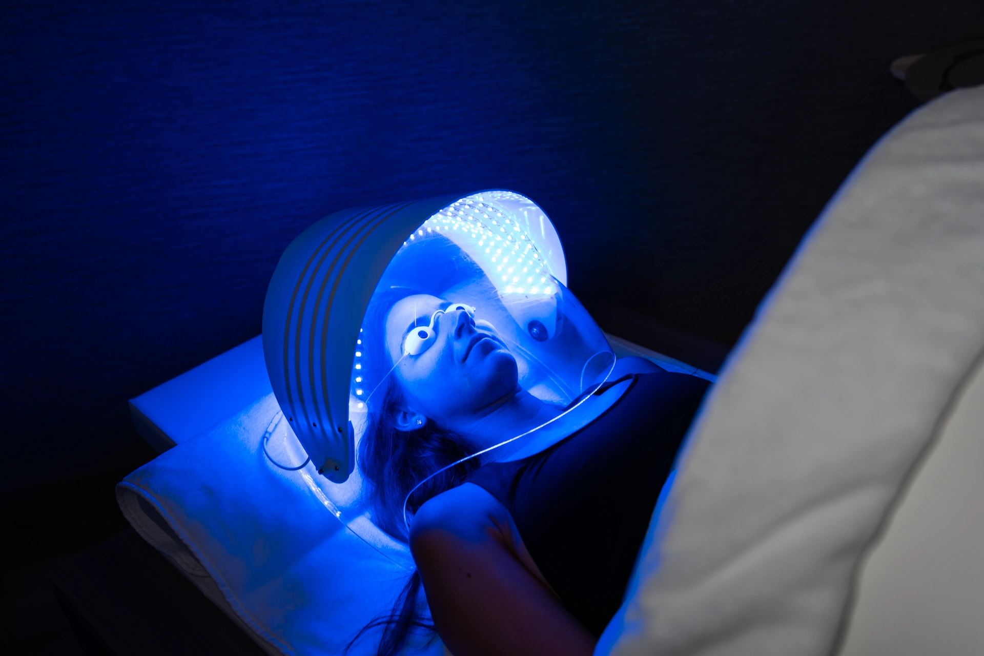 Person receiving LED light therapy, lying under curved blue light panel with protective goggles in spa-like setting.