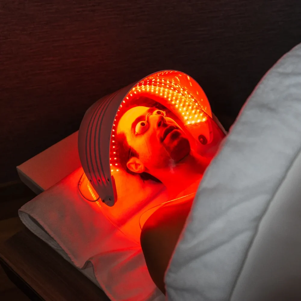 Person lying under curved red LED panel with goggles, receiving facial light therapy in calm wellness setting