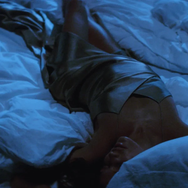 Guest resting diagonally across white bed in metallic dress, bathed in moody blue light and surrounded by soft textures