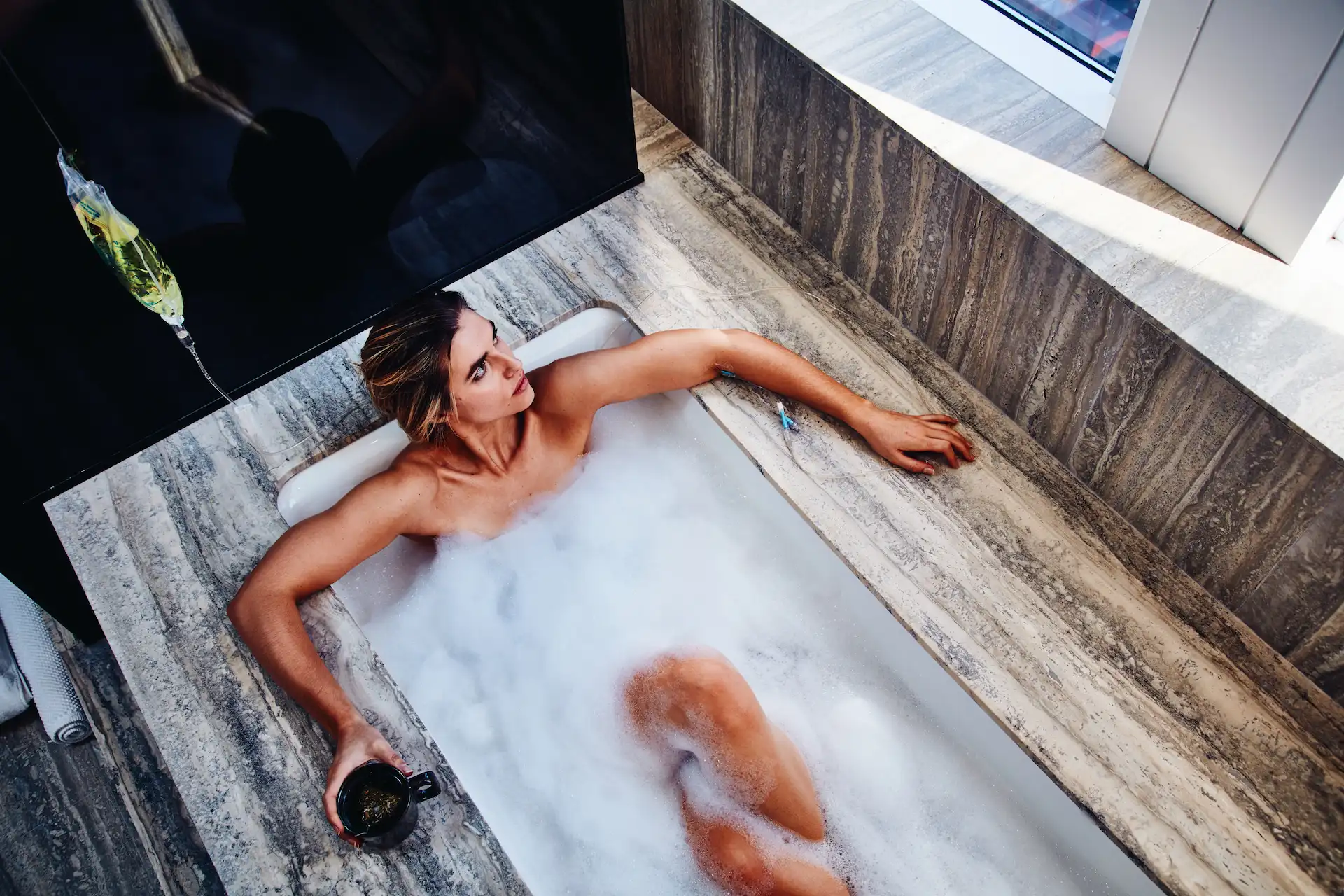 Person relaxing in foam-filled bathtub with dark cup in hand, natural light filtering into marble-tiled bathroom