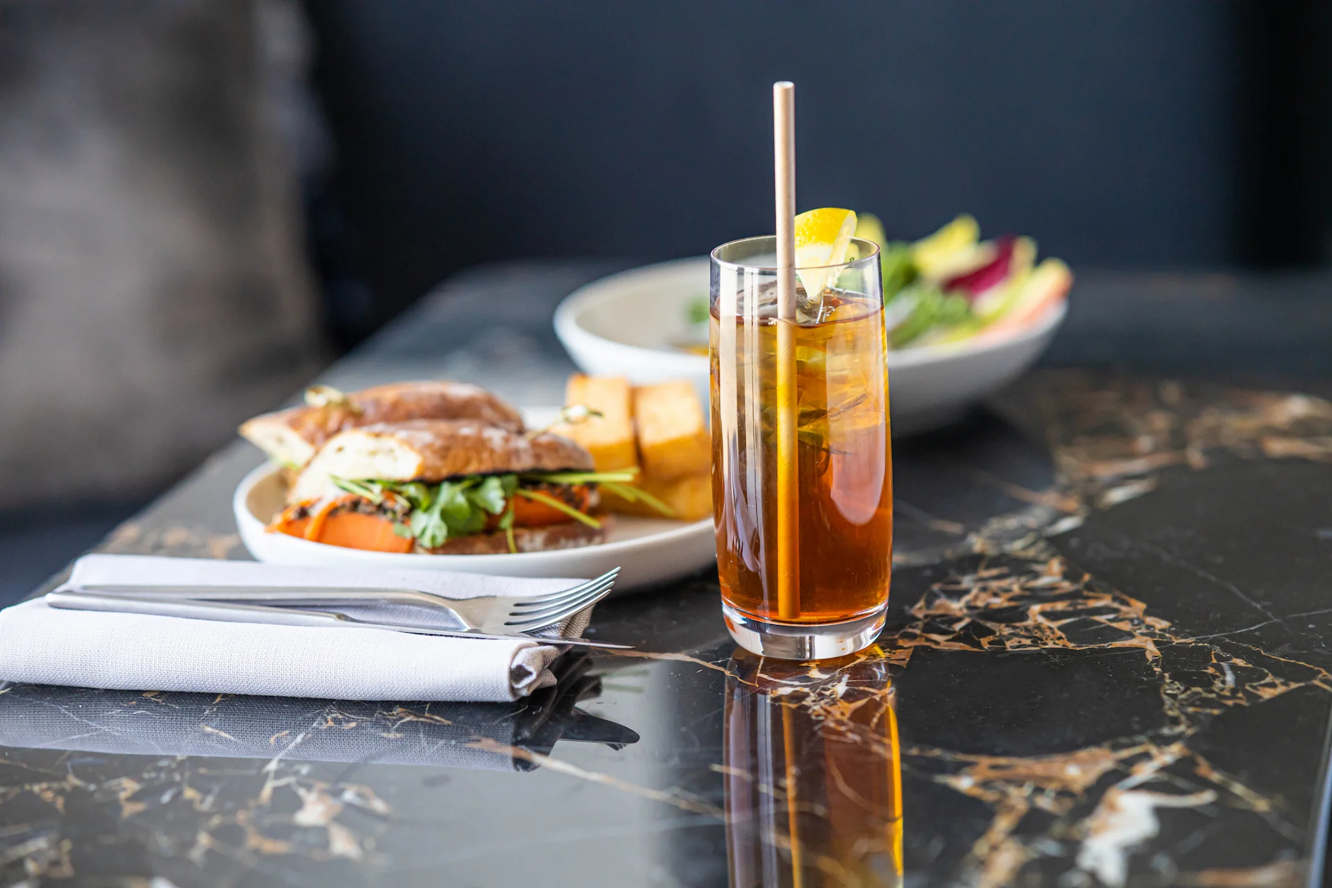Balanced meal with sandwich, potato wedges, salad, and iced tea served on dark marble table in refined dining setting