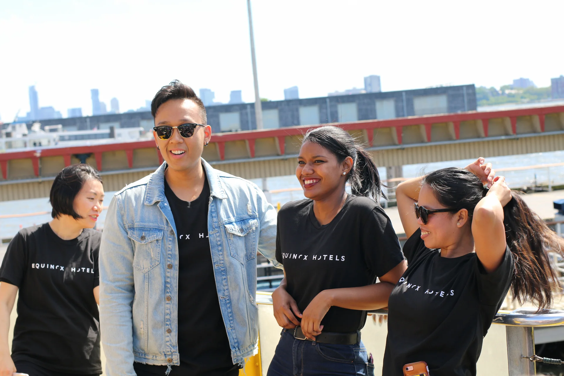 Equinox Hotels team members in branded shirts standing by waterfront with city skyline, sharing a casual moment outdoors