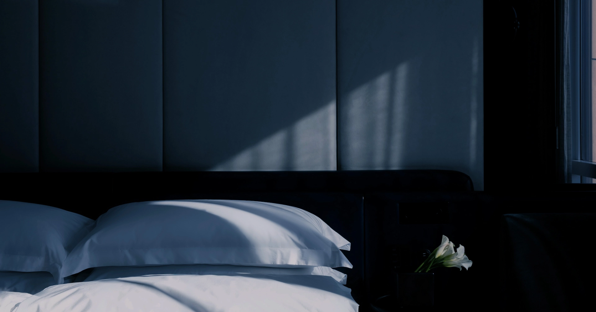 Tranquil hotel bedroom with soft white bedding, moody shadows, and white flowers adding a touch of quiet elegance