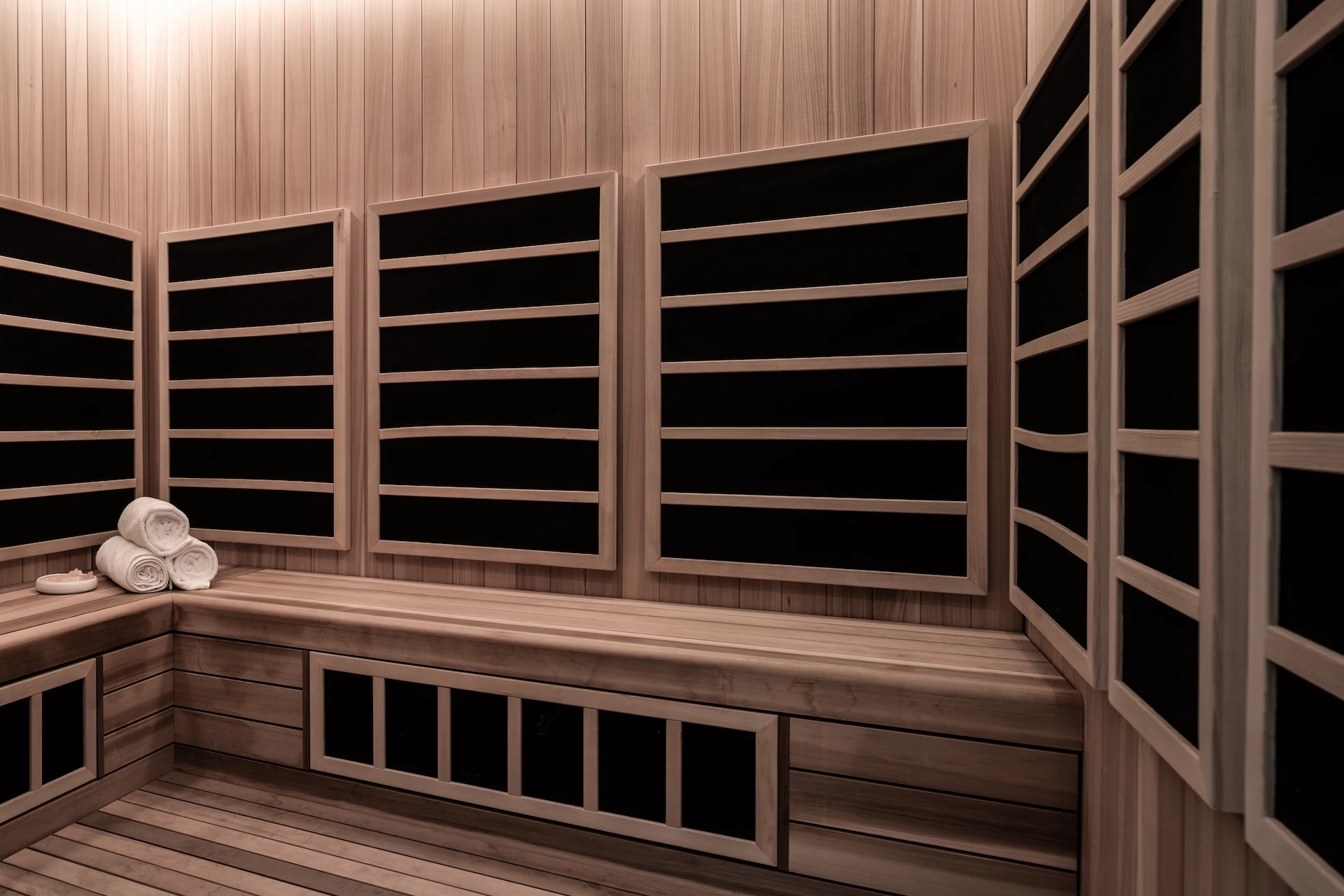 Interior of infrared sauna with wooden panels, black heaters, and rolled towels on bench, evoking minimalist wellness