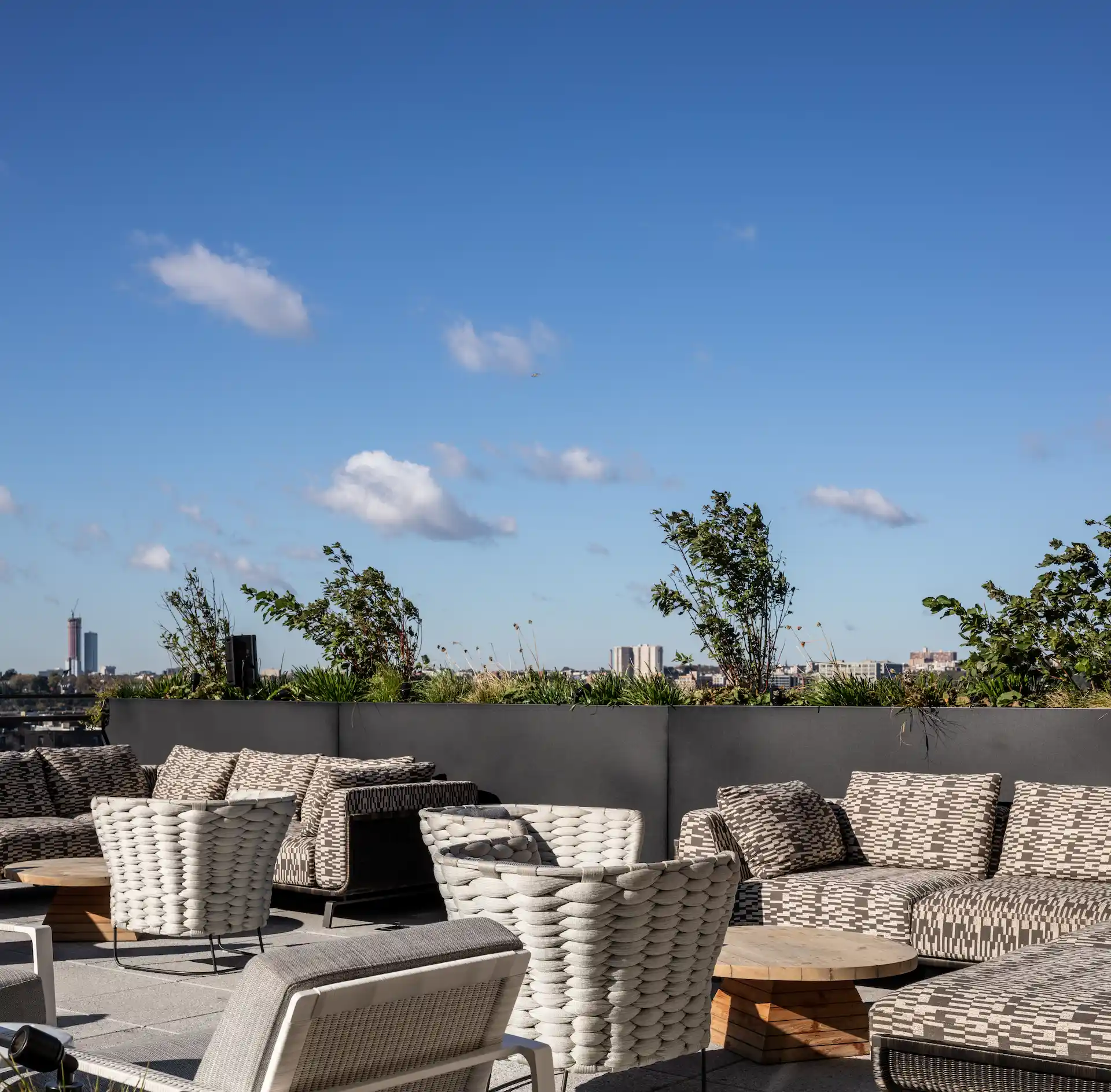 Rooftop lounge with woven chairs, greenery, and skyline views under blue sky
