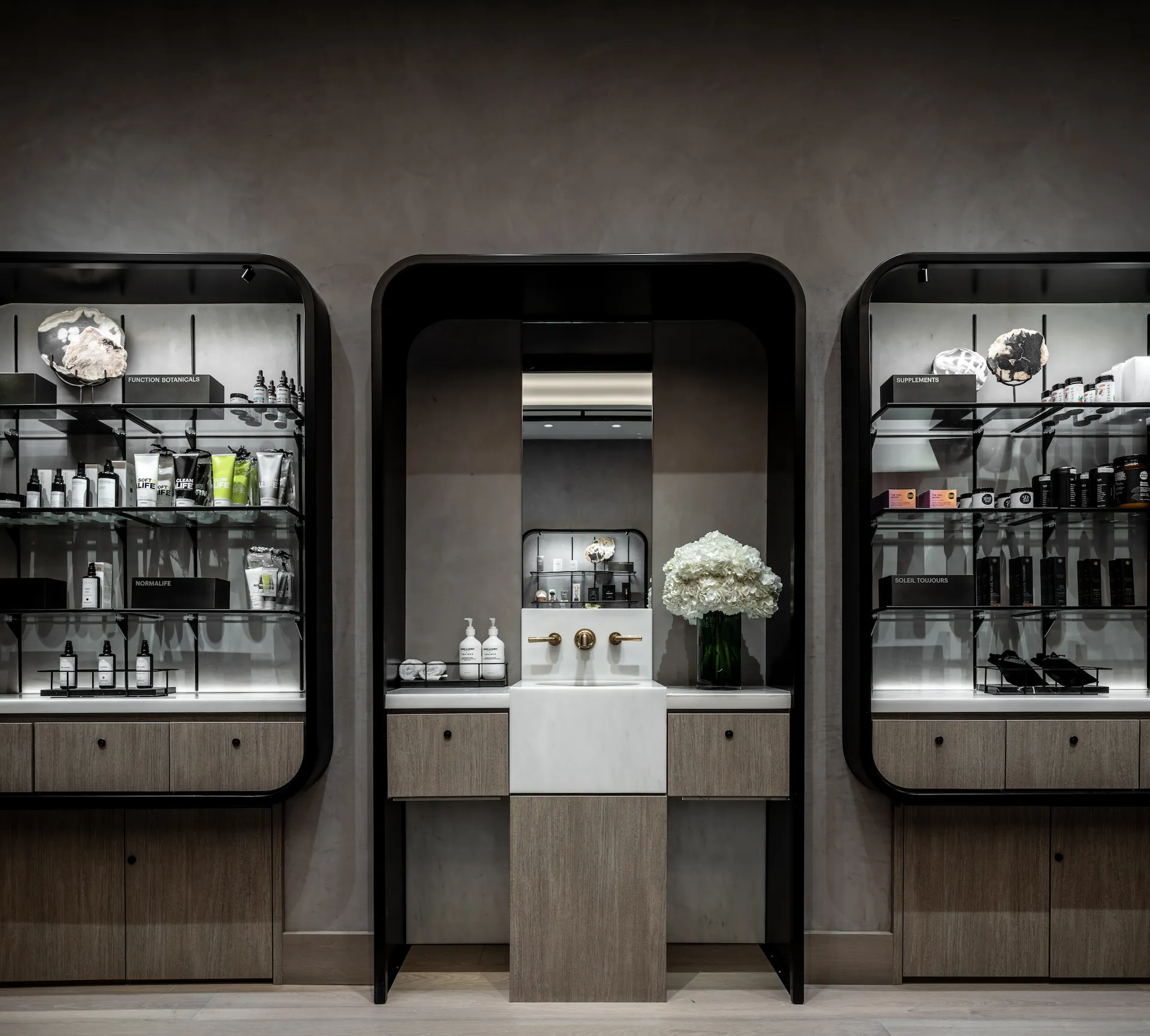 Elegant spa vanity with gold fixtures, white sink, and shelves displaying skincare products and white flowers in symmetrical layout