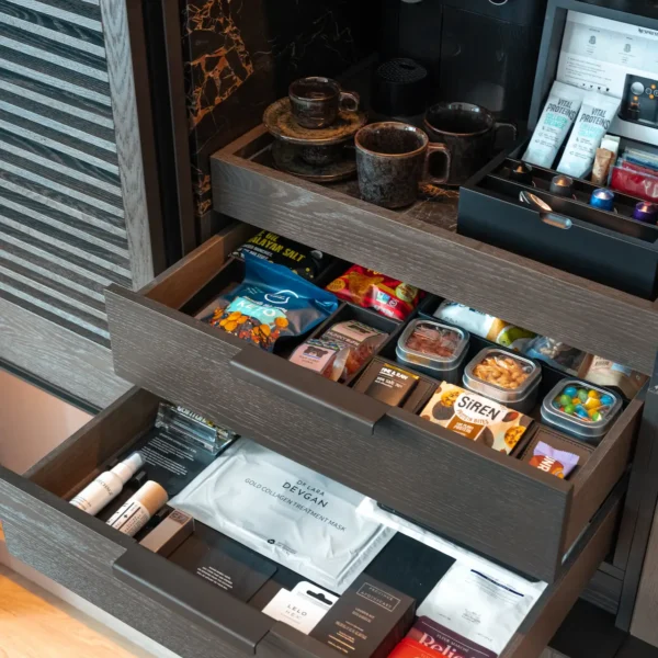 Open cabinet with curated minibar drawers featuring coffee setup, gourmet snacks, and personal care items, evoking tailored comfort and elevated hospitality