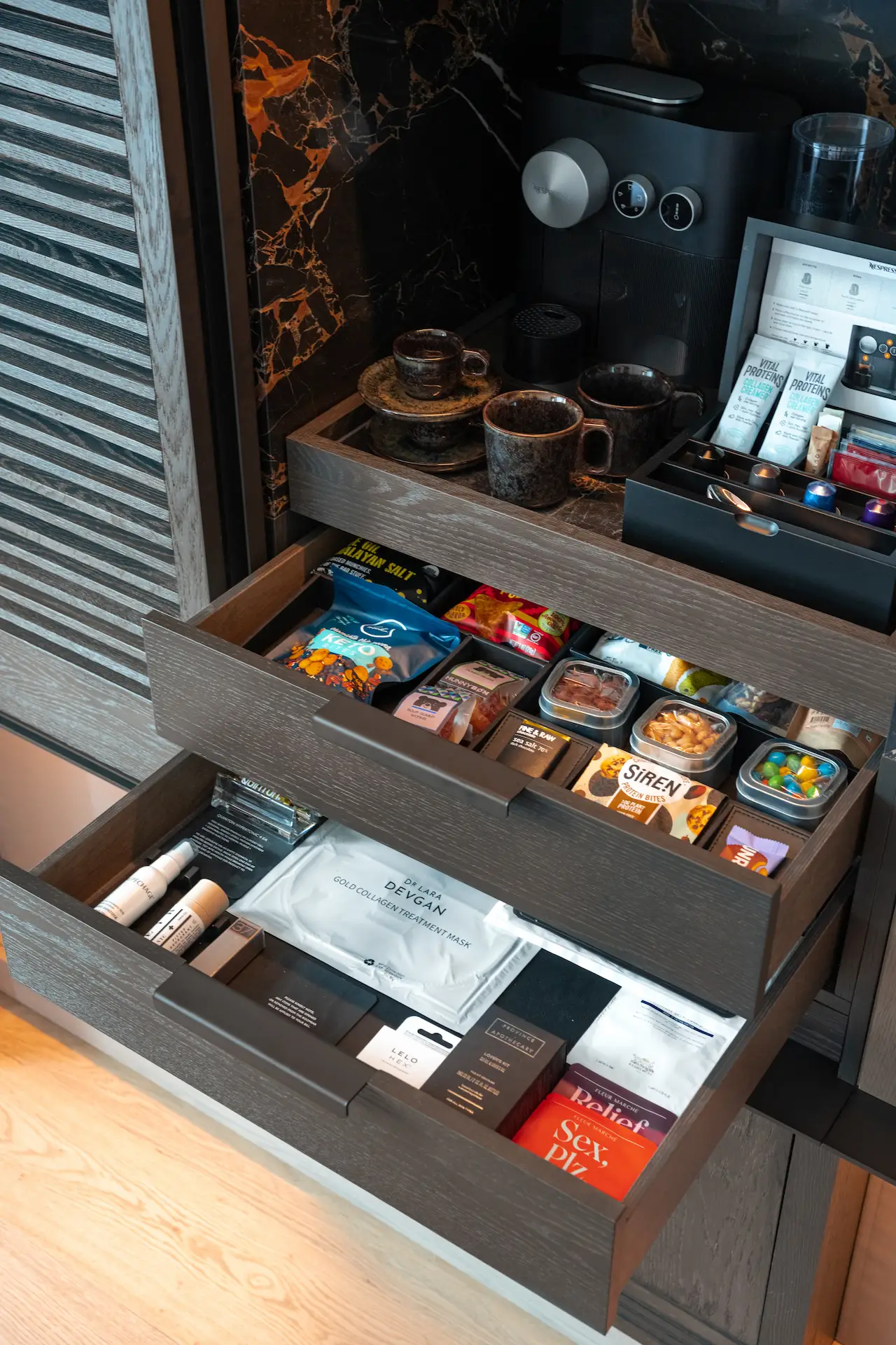 Open cabinet with curated minibar drawers featuring coffee setup, gourmet snacks, and personal care items, evoking tailored comfort and elevated hospitality