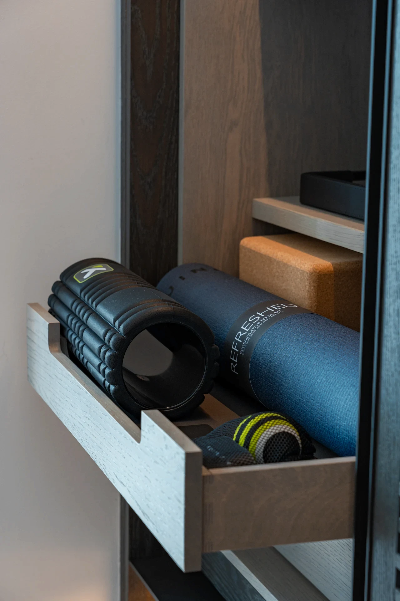 Drawer with foam roller, yoga mat labeled “REFRESH,” cork blocks, and resistance band neatly arranged in wooden cabinet