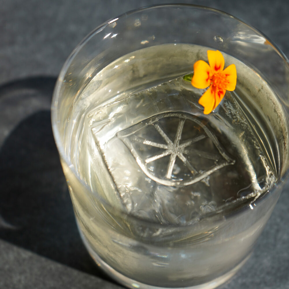 Glass with clear drink and patterned ice cube, topped with orange-yellow flower on dark surface.