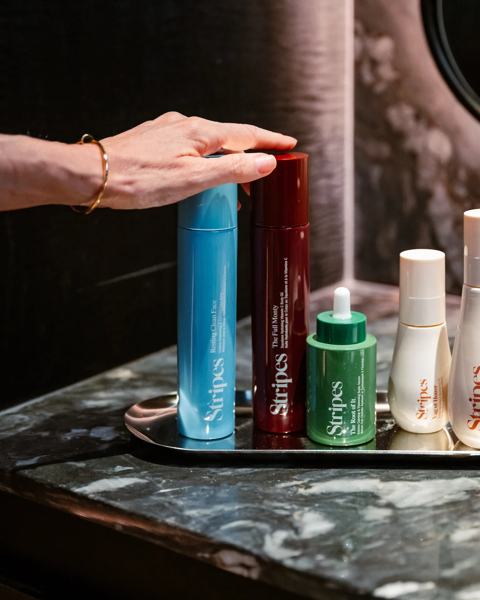 Hand reaching toward colorful Stripes skincare bottles on tray over dark marble countertop in vanity setting