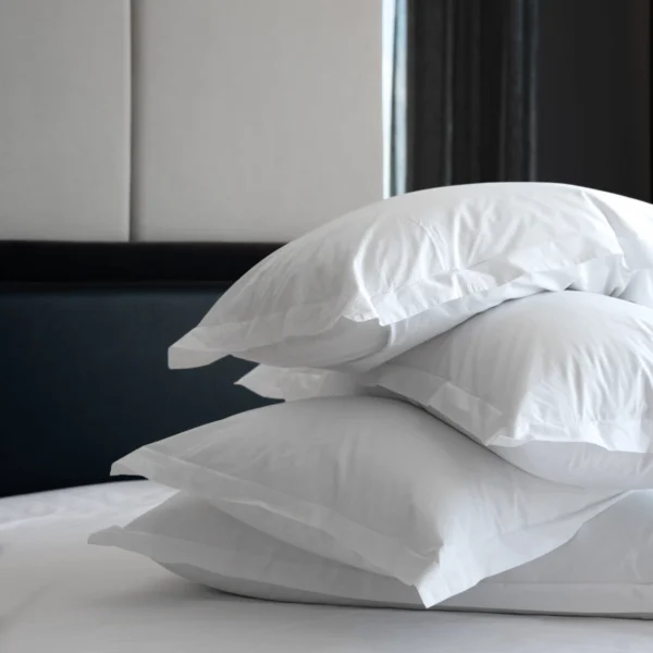 Stack of four white pillows on a white bed at Equinox Hotel with dark headboard & sheer curtained window in natural light.