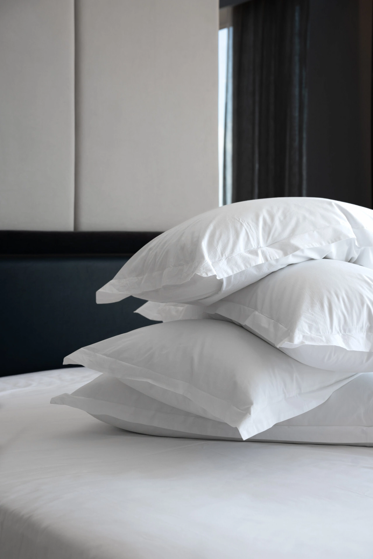 Stack of four white pillows on a white bed at Equinox Hotel with dark headboard & sheer curtained window in natural light.