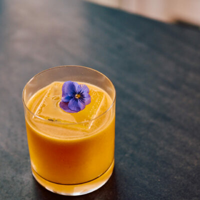 Glass of bright orange drink with ice cubes, topped with a purple edible flower on dark surface.