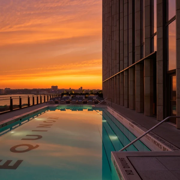 Rooftop pool at sunset with “EQUINOX” on pool floor, bordered by glass railing and city views, evoking serene luxury and elevated urban escape