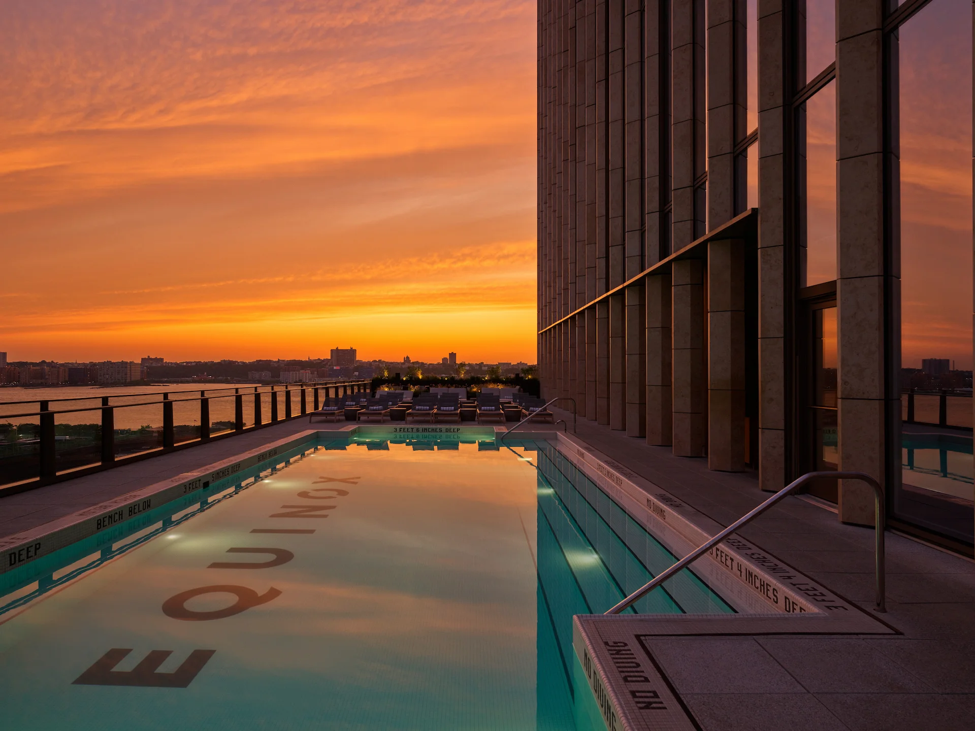 Rooftop pool at sunset with “EQUINOX” on pool floor, bordered by glass railing and city views, evoking serene luxury and elevated urban escape