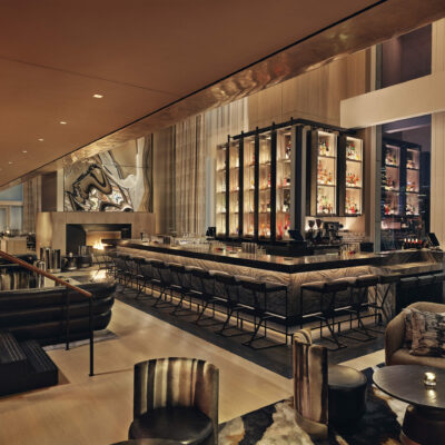 Luxurious bar with illuminated liquor shelves, high stools, plush seating, and city views through tall windows.