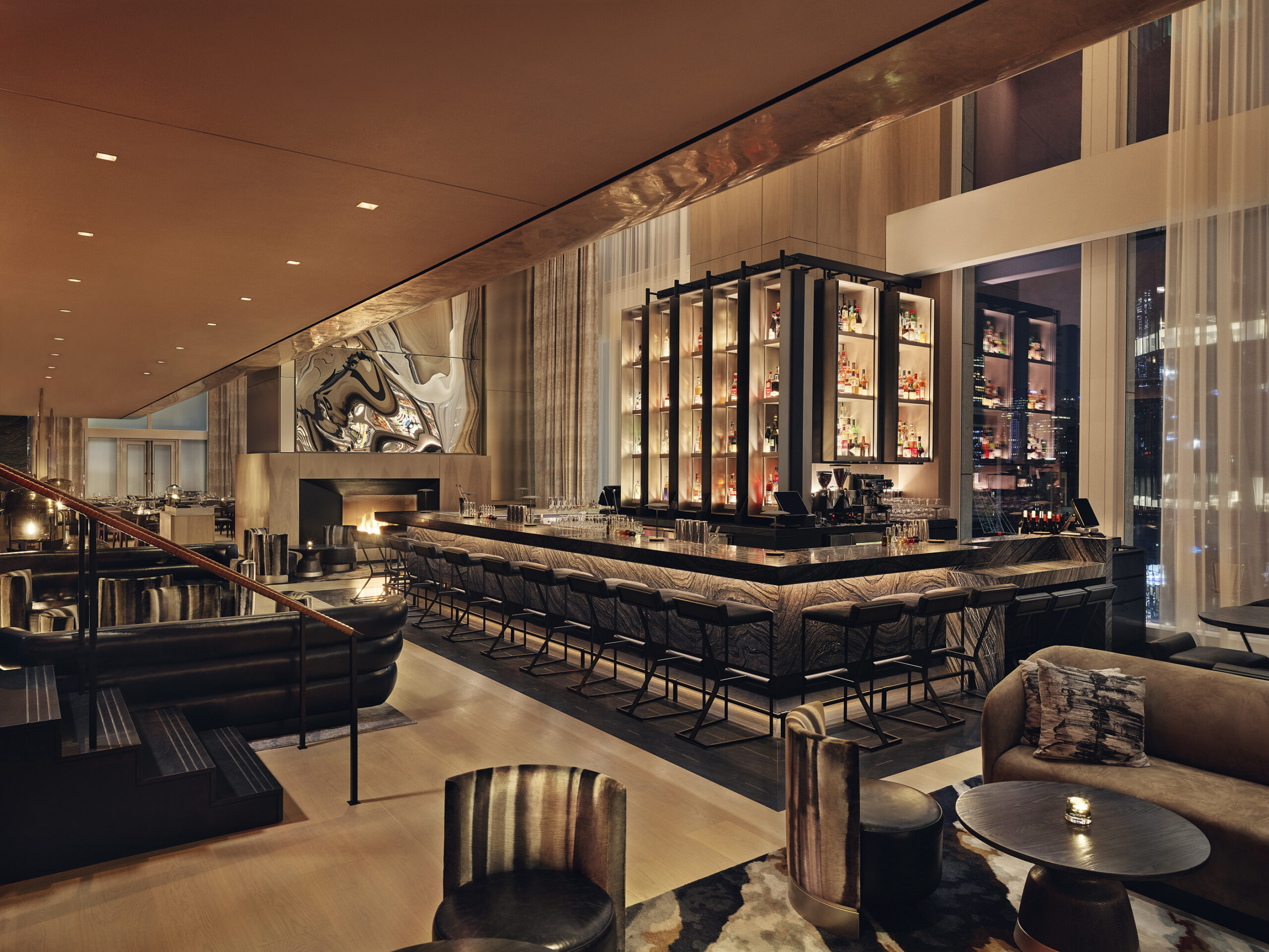 The bar at night, a large square counter with a lit bottle display at its centre, lounge chairs and low tables around it and a fireplace burning to one side.