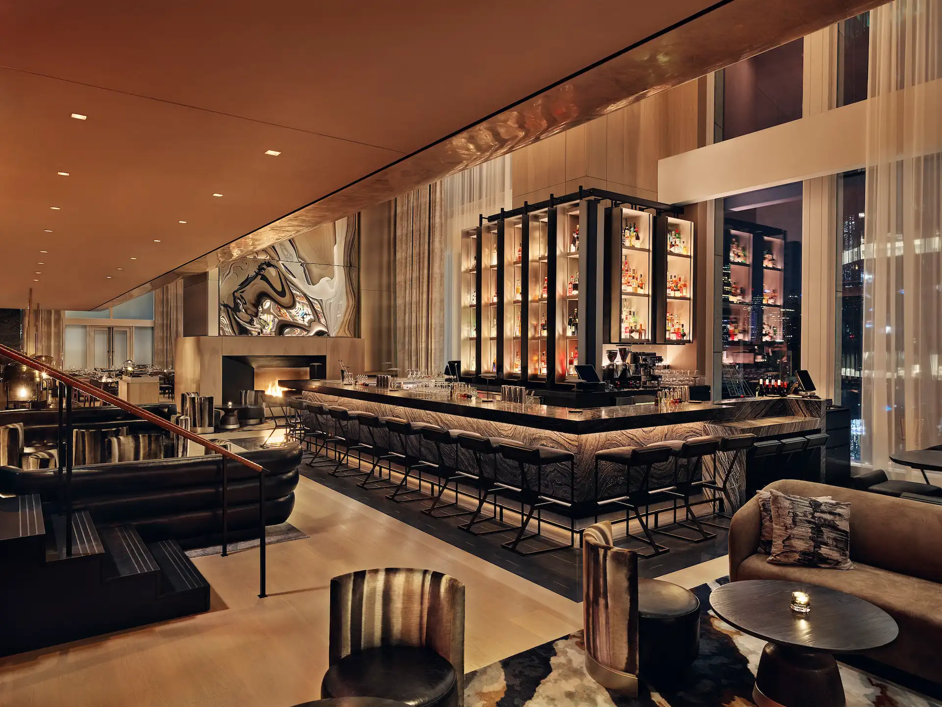 Modern bar lounge featuring a sleek marble counter, warm ambient lighting, plush seating, and sweeping nighttime city views, creating an inviting atmosphere of sophisticated urban luxury.
