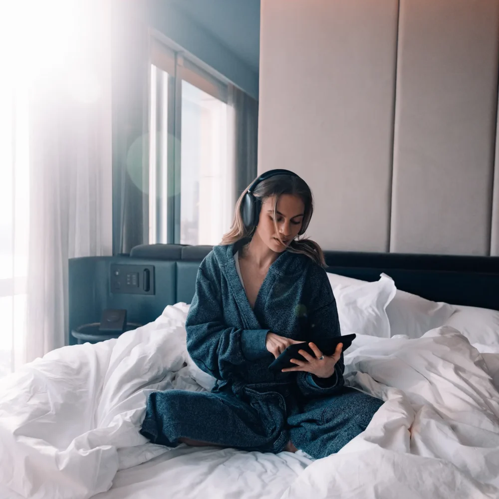 Person in dark robe with headphones using tablet on rumpled bed, bathed in soft sunlight in modern serene room