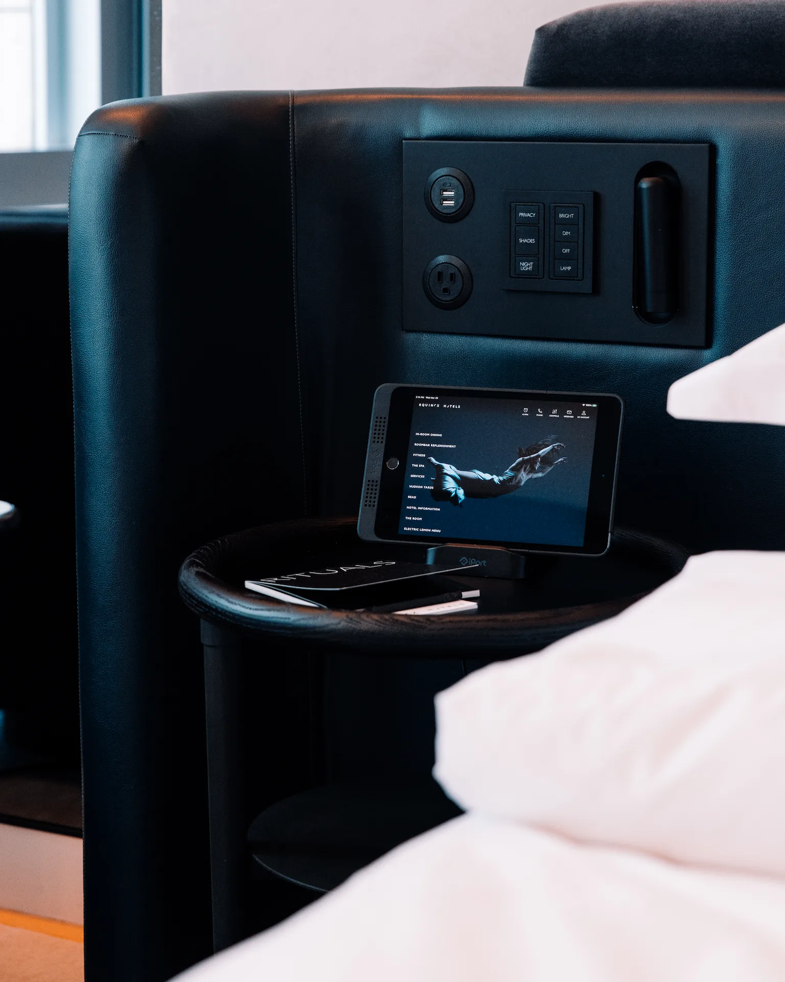 Smartphone on bedside table showing room controls interface, in sleek hotel room with black headboard and white linens