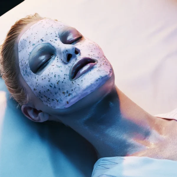 Person lying down with white speckled facial mask, eyes closed under bluish light in serene spa or skincare treatment setting