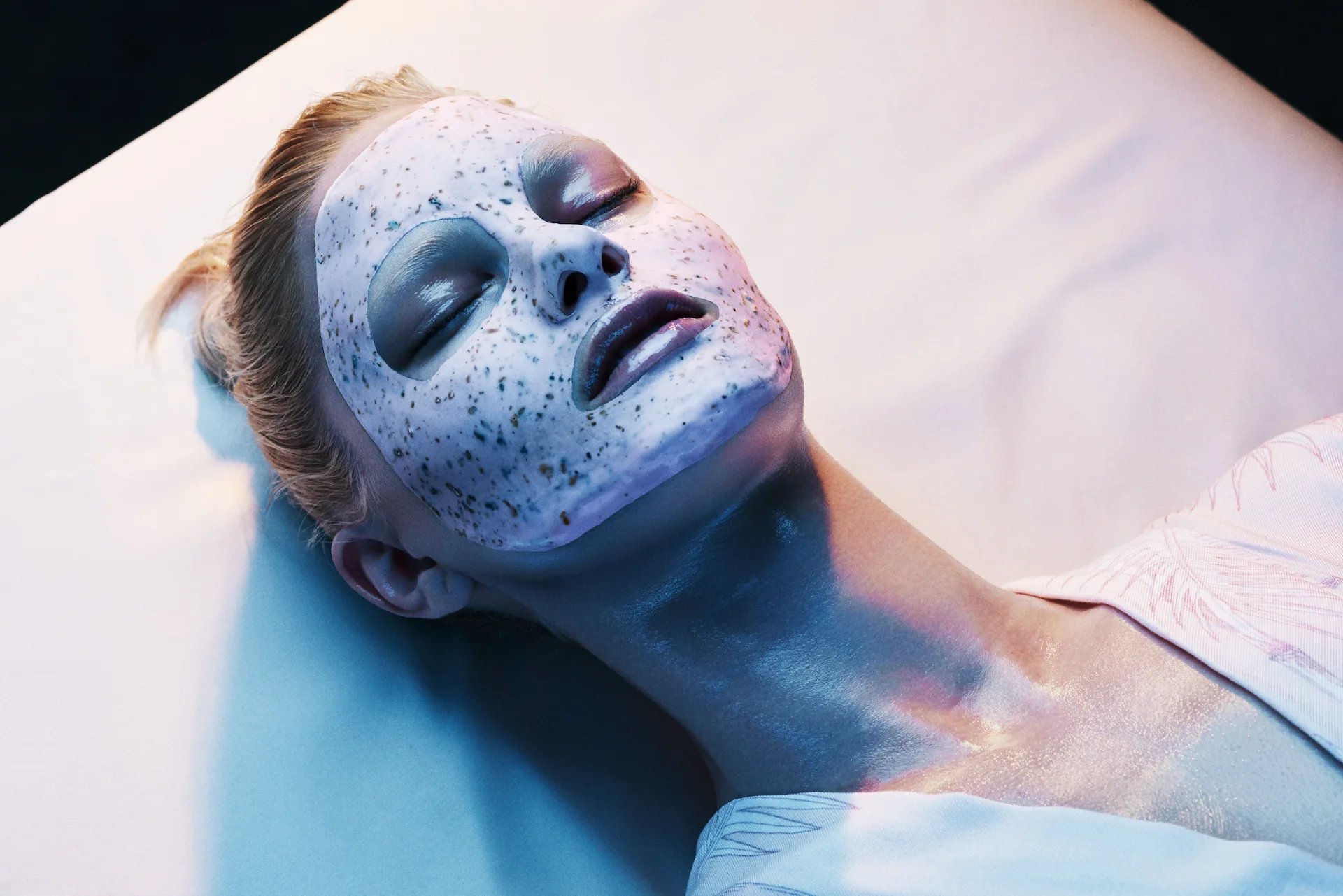 Person lying down with white speckled facial mask, eyes closed under bluish light in serene spa or skincare treatment setting