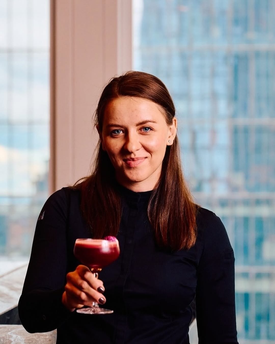 Person holding a cocktail with dark red foam, standing indoors near large windows overlooking city skyscrapers.