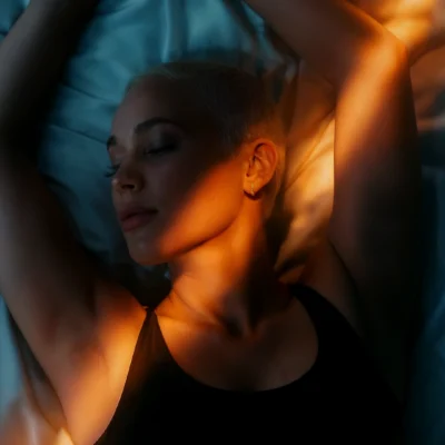 Person lying on bed with arms stretched overhead, warm orange light streaks across face and body contrasting with cool blue sheets