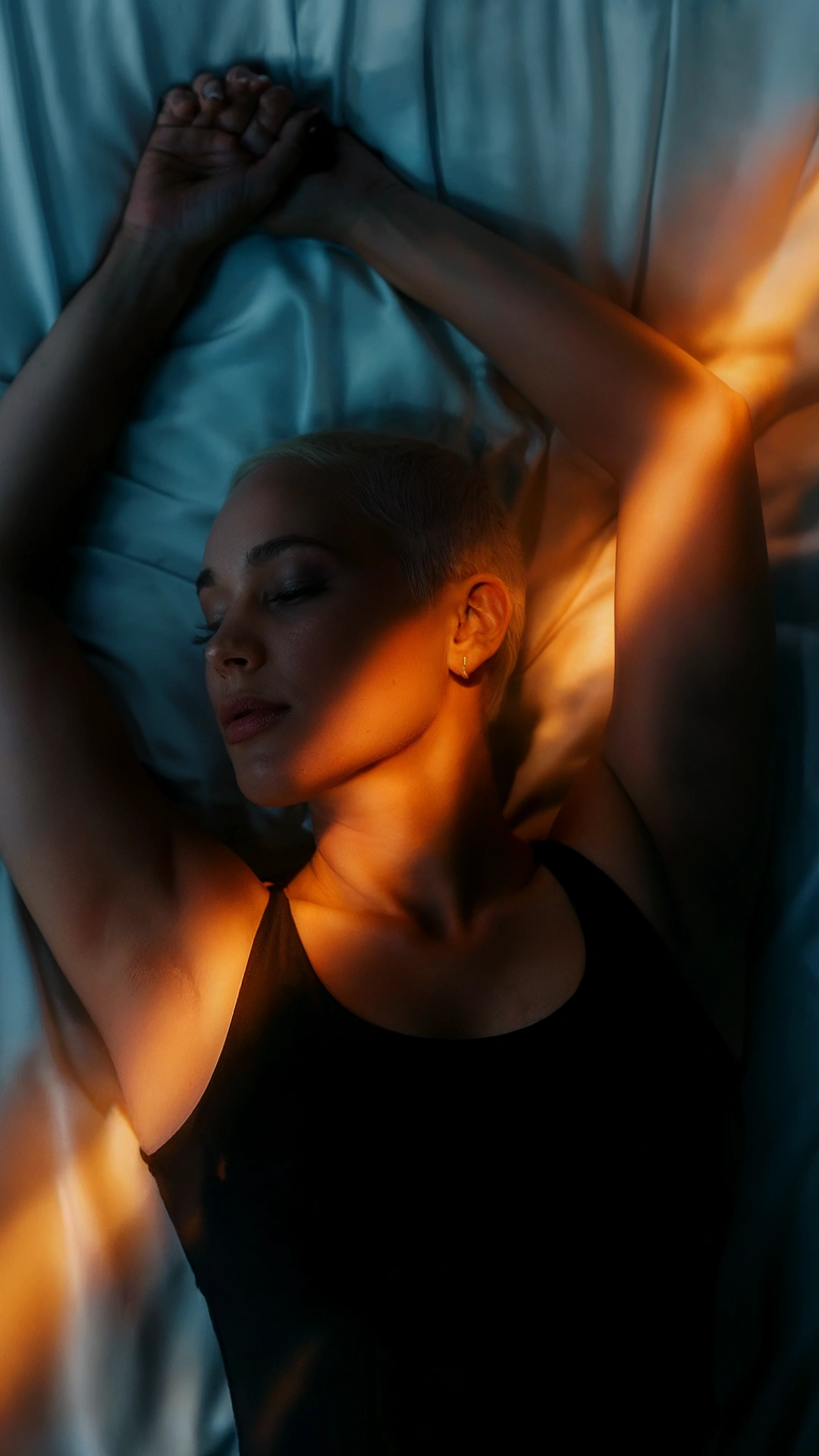 Person lying on bed with arms stretched overhead, warm orange light streaks across face and body contrasting with cool blue sheets
