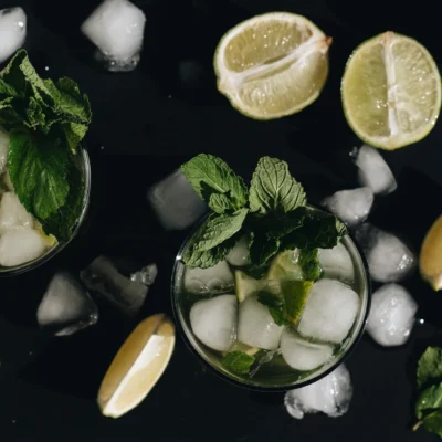 Flat lay of two mint-lime drinks with ice, limes, and mint leaves on dark surface.
