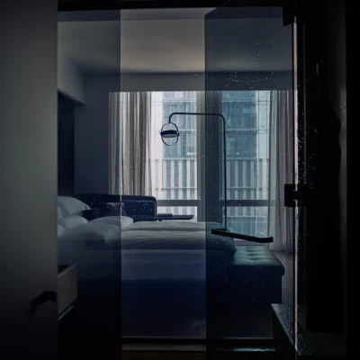 Dimly lit modern hotel bedroom viewed through a glass partition, featuring plush white linens, a dark elegant headboard framed by sheer curtains, and a breathtaking nighttime cityscape beyond, creating an intimate, luxurious urban retreat.