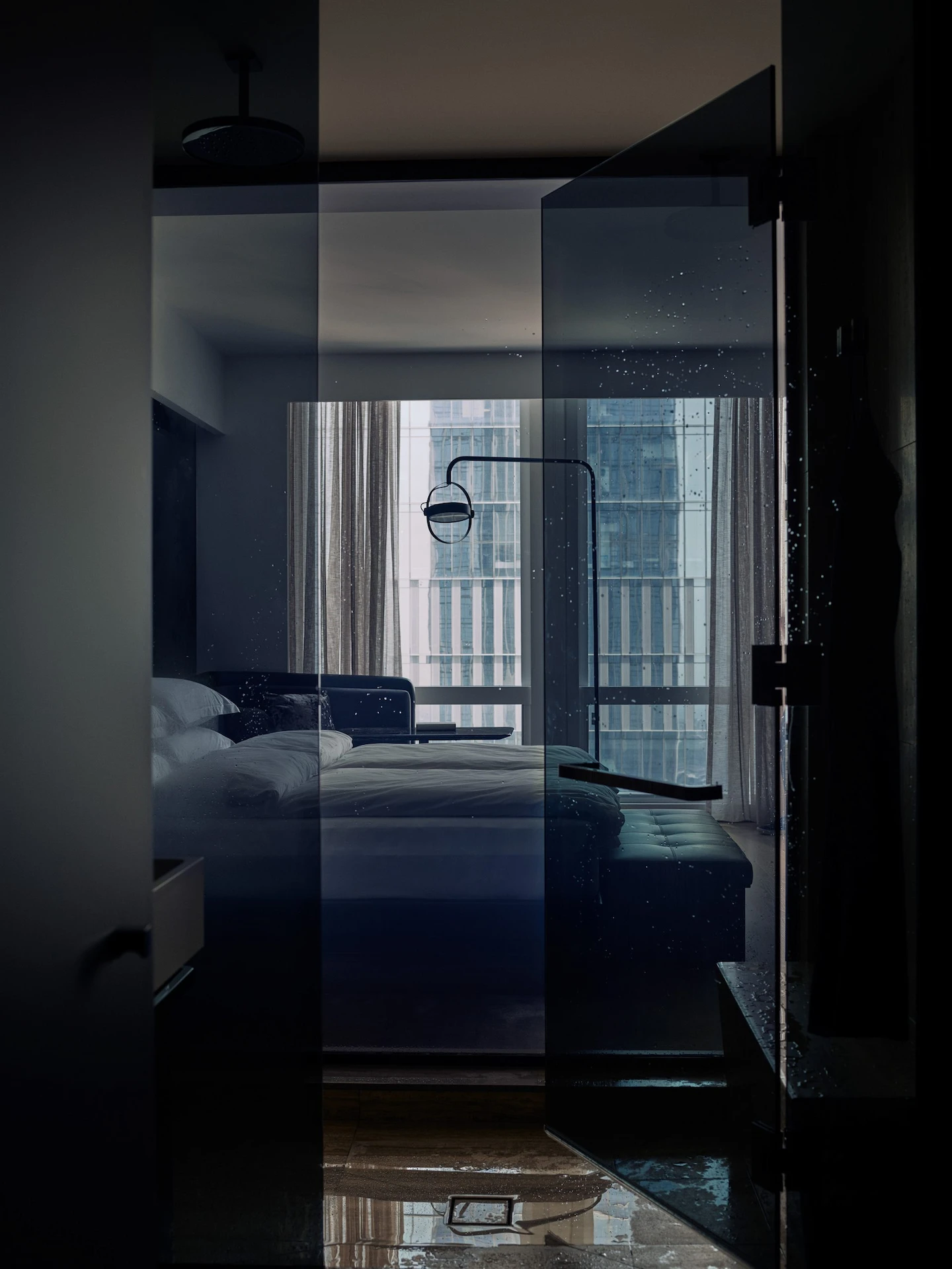 Dimly lit modern hotel bedroom viewed through a glass partition, featuring plush white linens, a dark elegant headboard framed by sheer curtains, and a breathtaking nighttime cityscape beyond, creating an intimate, luxurious urban retreat.