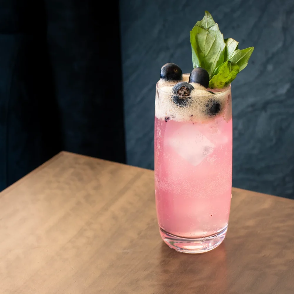 Tall glass of pink drink with blueberries and basil on wooden surface, frothy top and dark background enhancing contrast