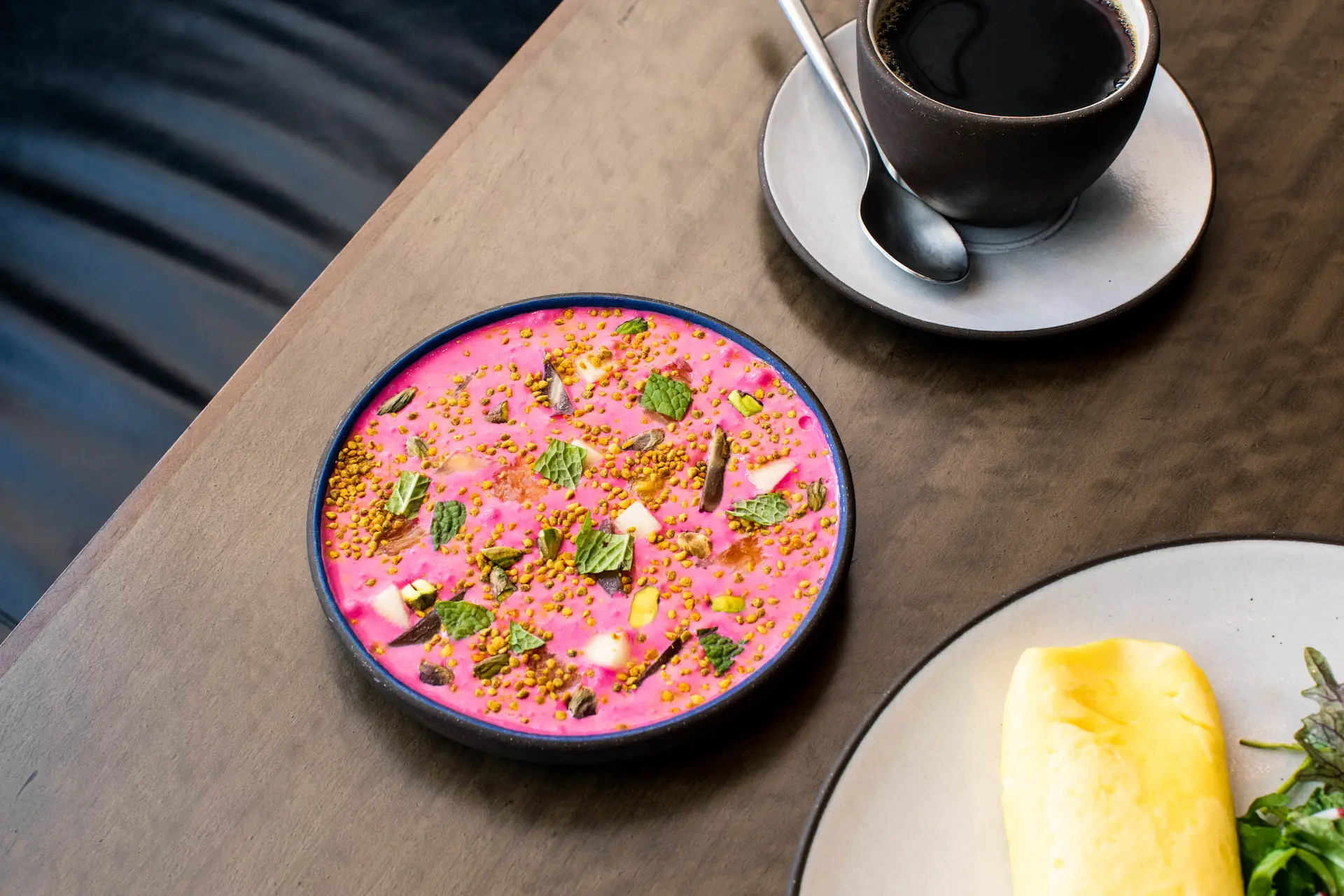 Pink smoothie bowl with mint and granola, black coffee, and salad plate on wooden table