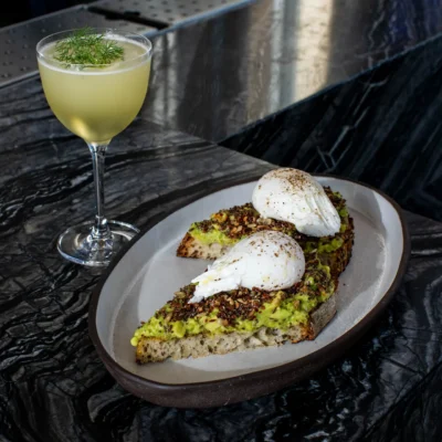 Avocado toast topped with perfectly poached eggs, accompanied by a pale cocktail on a marbled surface, capturing a refined brunch moment with elegant presentation and vibrant textures.