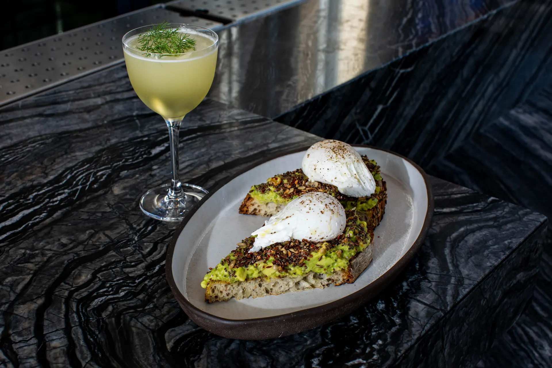 Avocado toast topped with perfectly poached eggs, accompanied by a pale cocktail on a marbled surface, capturing a refined brunch moment with elegant presentation and vibrant textures.
