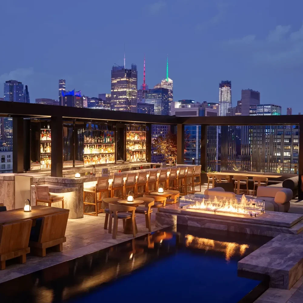 Rooftop bar at night with pool, fire pit, and city skyline, evoking vibrant urban luxury