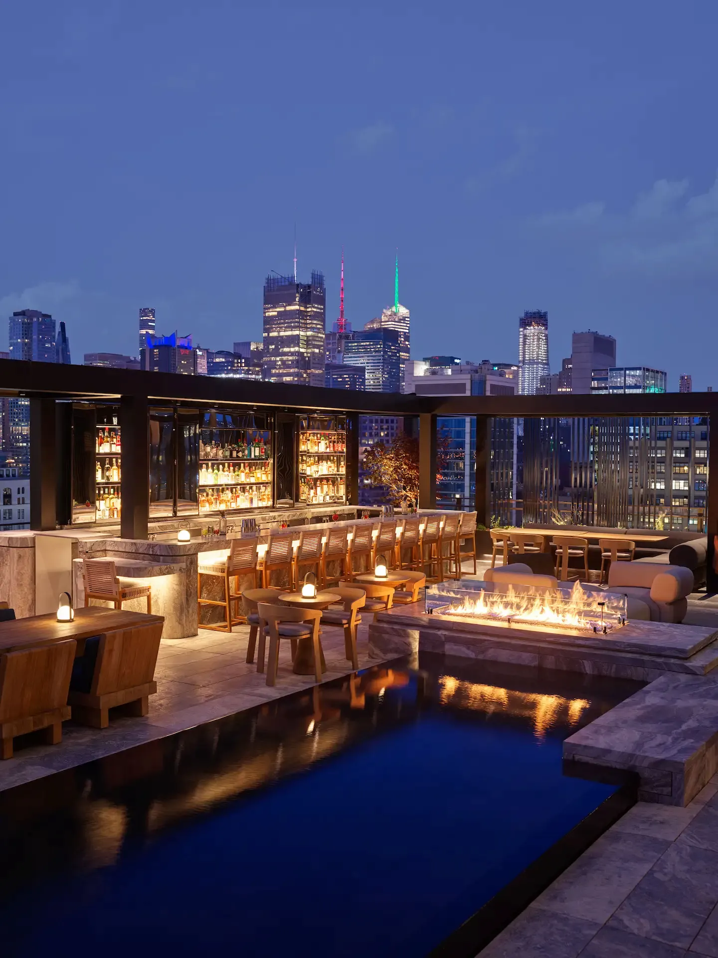 Rooftop bar at night with pool, fire pit, and city skyline, evoking vibrant urban luxury